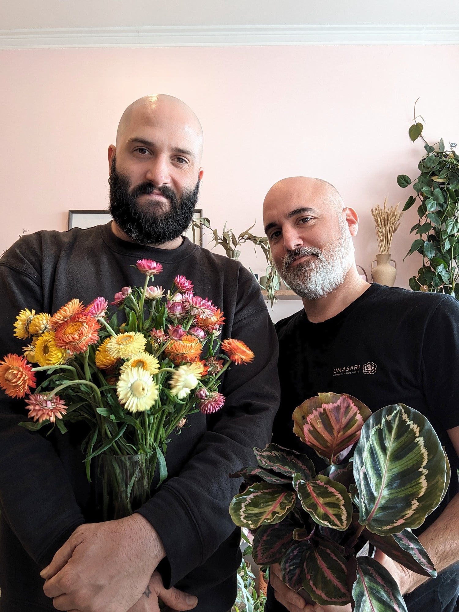 Two men standing side by side holding vibrant plants. The man on the left is holding a bouquet of colourful native Australian paper daisies in shades of yellow, orange, and pink, while the man on the right holds a lush green Calathea plant with variegated leaves. Both men have beards and are smiling softly, with a background of light pink walls and more plants behind them, creating a welcoming and natural florist environment