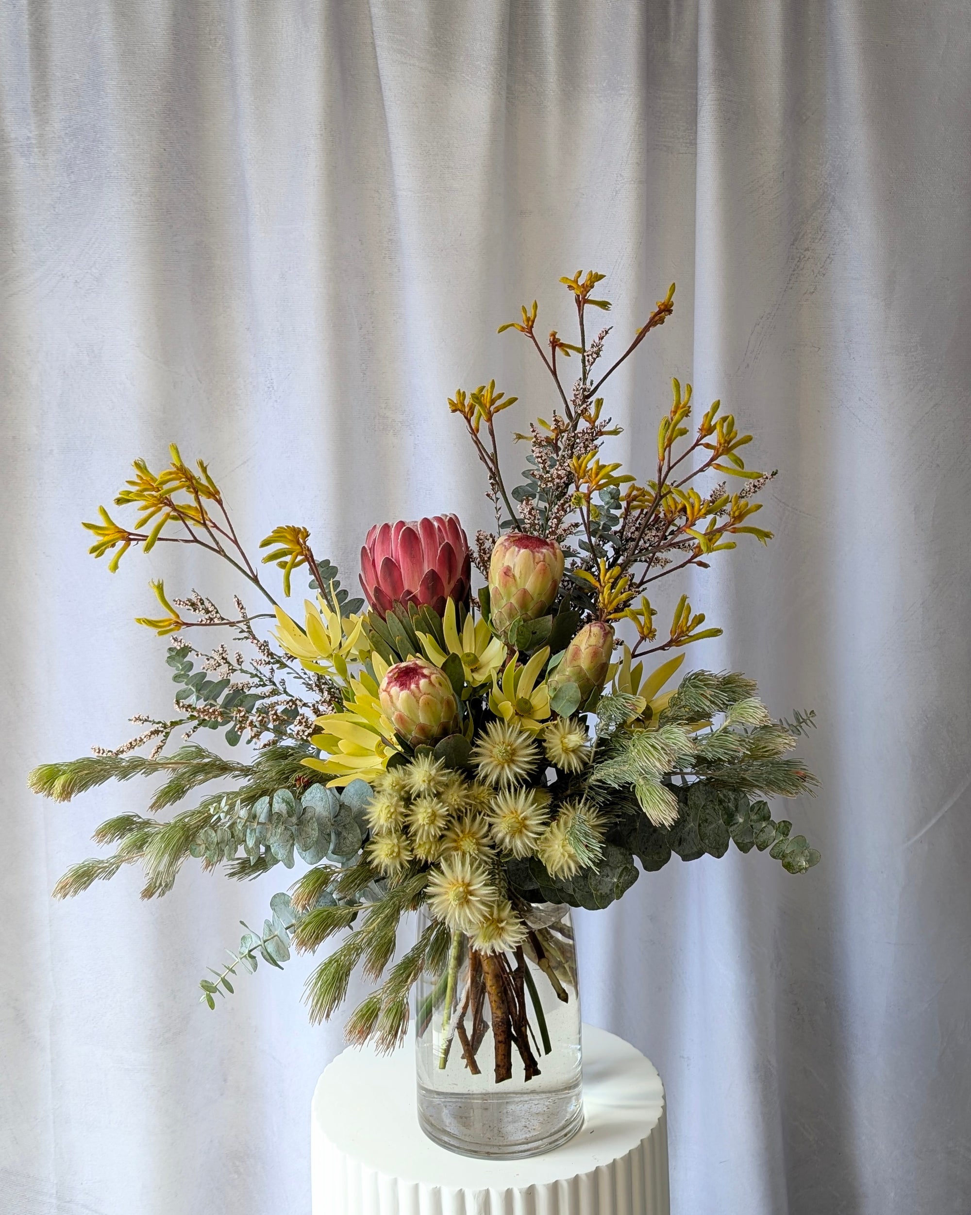 Bouquet of Australian Native flowers in a clear vase on a white pedestal against a white curtain background for delivery in Melbourne