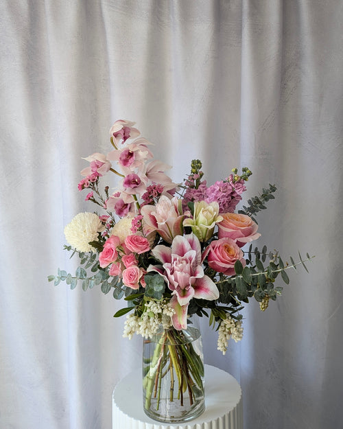 Bouquet of luxury flowers in a clear vase on a white surface with a light grey curtain background. Flowers include orchids, lilies, roses and chrysanthemums. Available for delivery in Melbourne. 