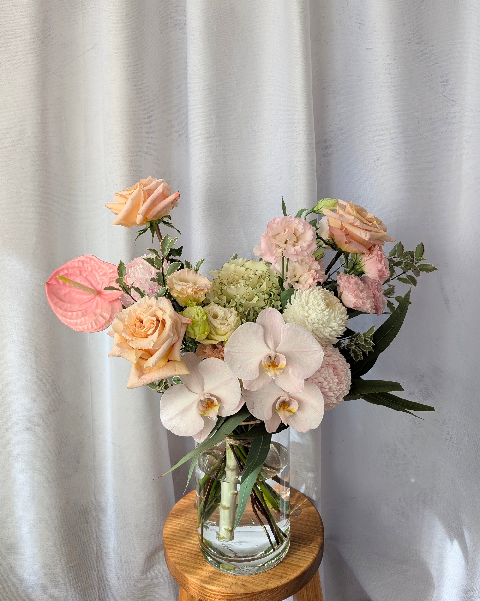 Bouquet of flowers  including orchids, anthuriums and roses in a clear vase on a wooden table with a white curtain background. For delivery in Melbourne