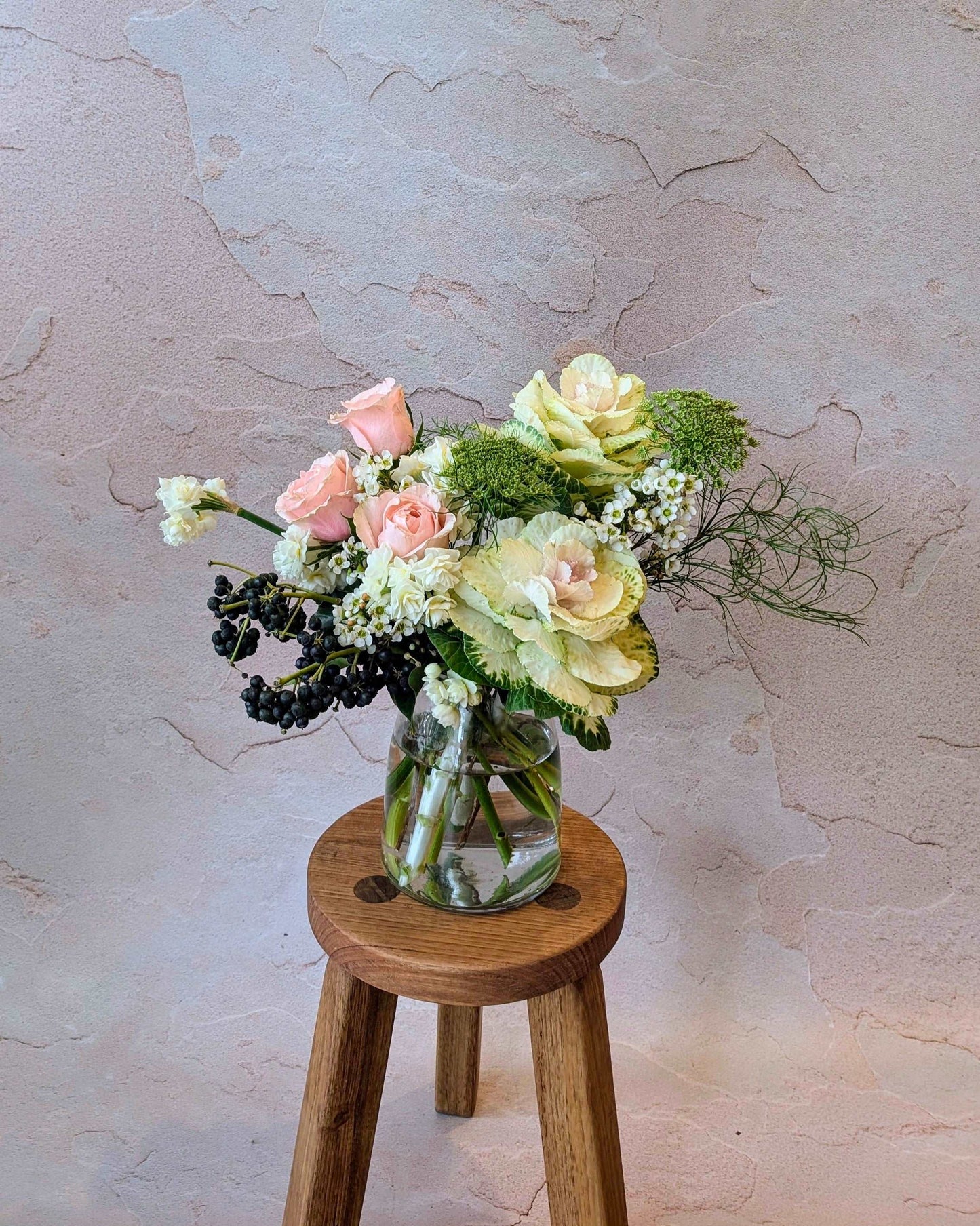 Petite floral arrangement featuring pale pink roses, ornamental cabbage, wax flowers, and mixed greenery in a clear vase on a wooden stool. Ideal for intimate floral decor, small gifts, or table centerpieces. The soft pastel tones create a gentle, elegant look