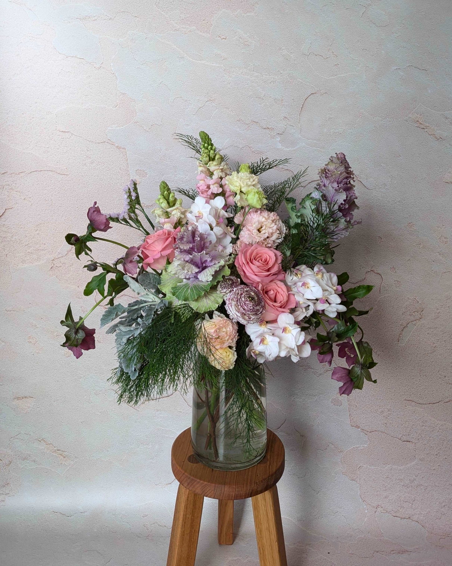 This image features a beautiful floral arrangement in a clear glass vase, placed on a wooden stool. The bouquet includes a mix of delicate pink roses, light peach lisianthus, delicate white orchis, purple kale, complemented by lush greenery such as dusty miller and fern-like foliage. Soft lavender and mauve flowers are artfully scattered throughout, adding depth and texture. The backdrop is a neutral-toned, textured wall, which enhances the elegance of the arrangement. This image would work well 