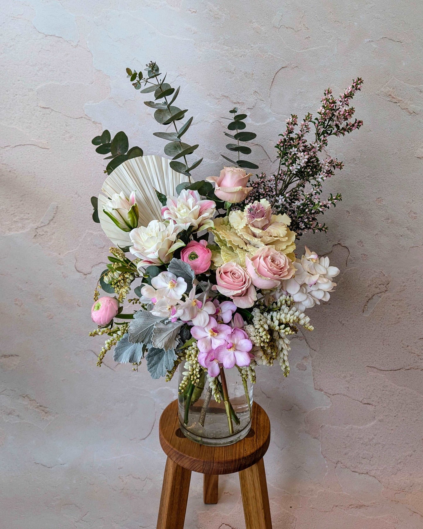 Soft pastel floral arrangement featuring pink roses, ranunculus, orchids, and mixed foliage with a dried palm fan in a clear vase on a wooden stool. Perfect for elegant decor, weddings, or romantic gifts. The muted pinks and creamy whites create a delicate, sophisticated aesthetic.