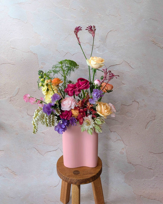 Chic floral arrangement in a pastel pink vase featuring roses, ranunculus, snapdragons, sweet peas, and kangaroo paw on a wooden stool. Ideal for modern home decor, boutique floral displays, and luxury gifts. Vibrant, colourful blooms create a stylish, contemporary aesthetic