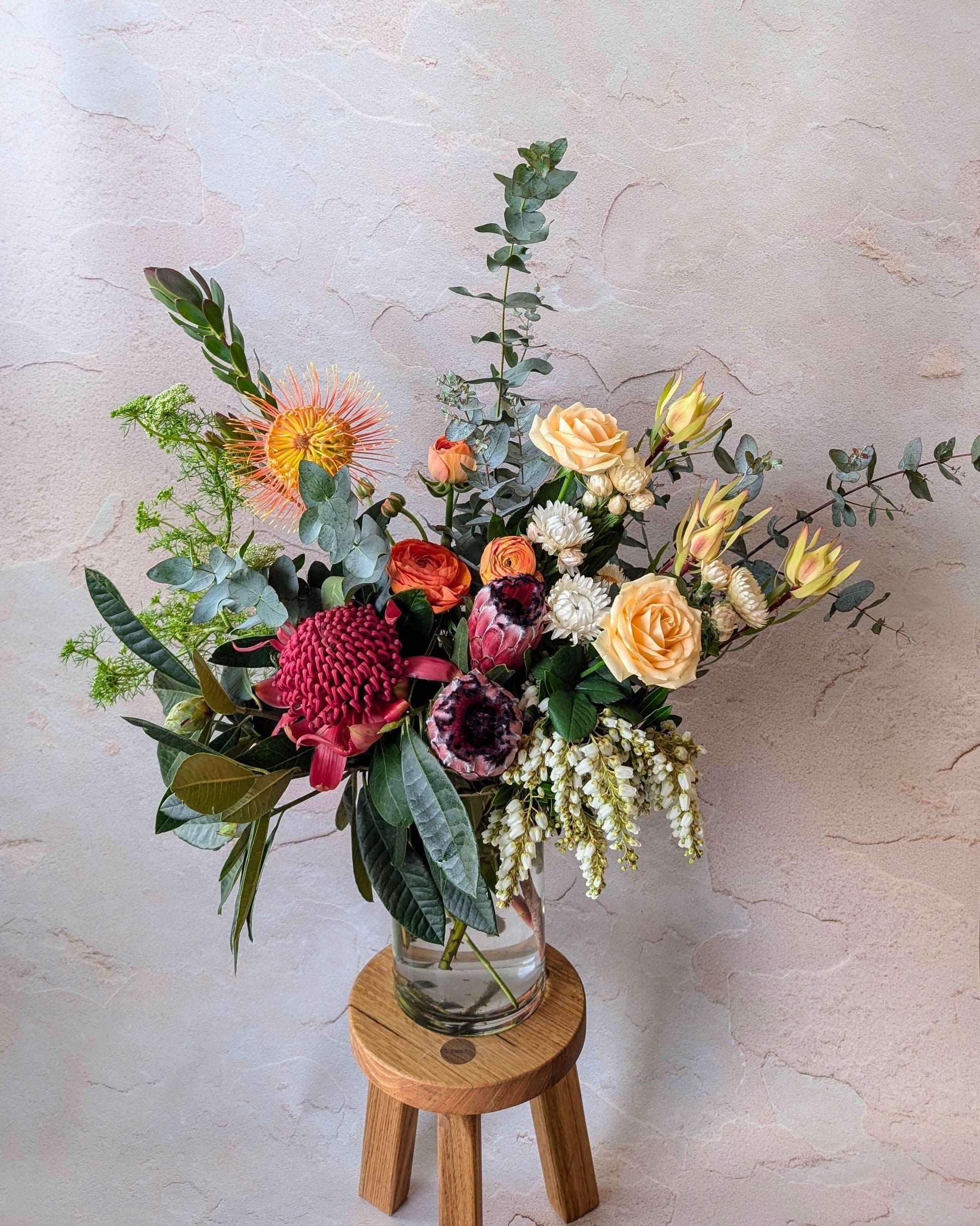 Elegant bouquet featuring vibrant native Australian flowers, including proteas, leucadendrons, roses, and eucalyptus leaves in a clear vase on a wooden stool. Perfect for modern floral arrangements, weddings, and interior decor. Boho-inspired floral design with fresh blooms and greenery.