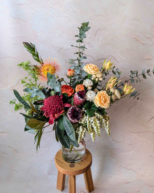 Elegant bouquet featuring vibrant native Australian flowers, including proteas, leucadendrons, roses, and eucalyptus leaves in a clear vase on a wooden stool. Perfect for modern floral arrangements, weddings, and interior decor. Boho-inspired floral design with fresh blooms and greenery.