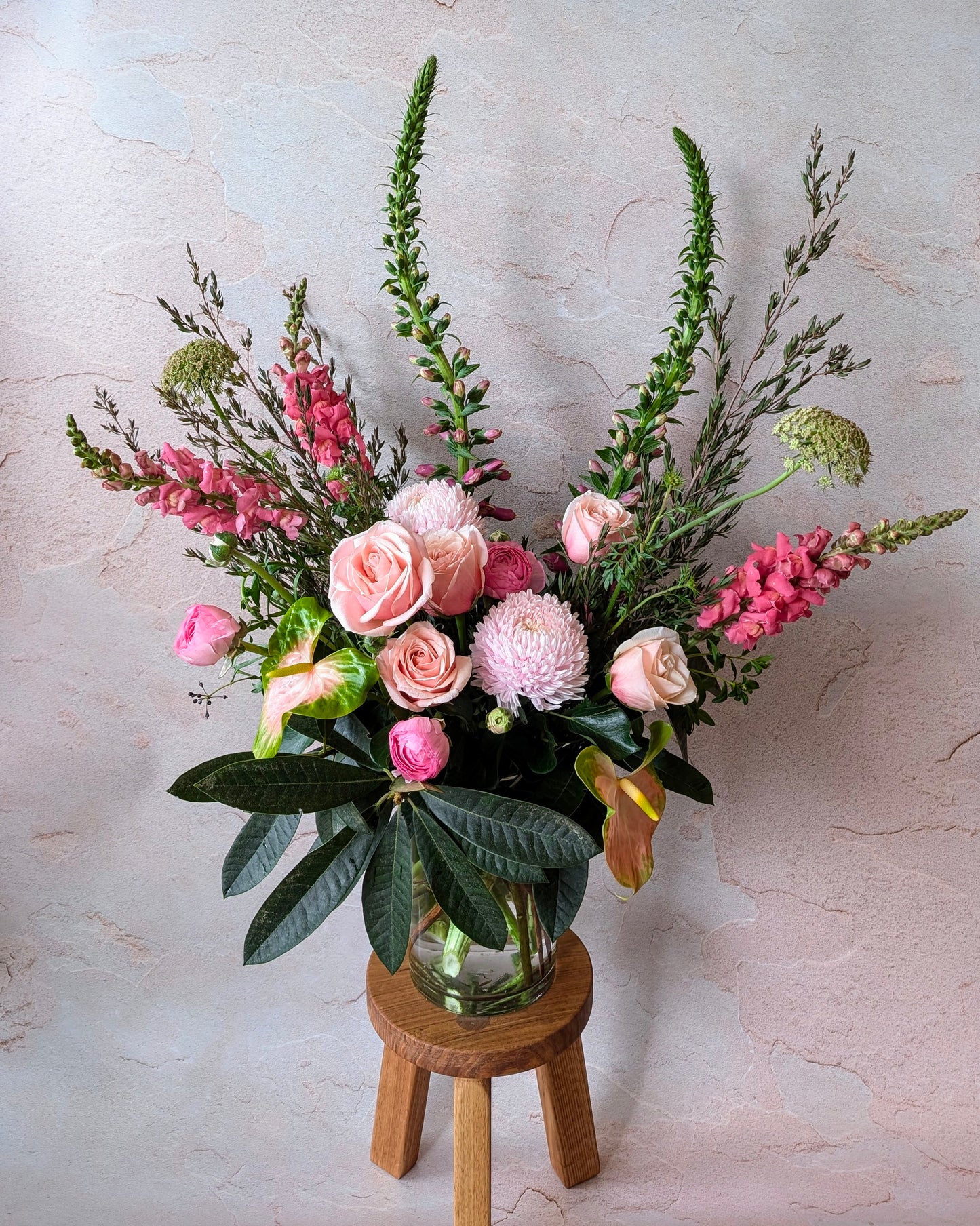 Bespoke pink floral arrangement with roses, chrysanthemums, and lush greenery in a clear vase on a wooden stool, designed by Umasari Florist, Thornbury, Melbourne. Perfect for weddings, special occasions, or gifting in Melbourne’s northern suburbs.