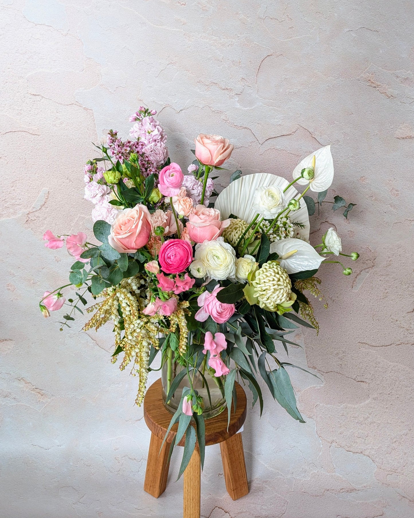 "Elegant pastel floral arrangement with roses, ranunculus, anthuriums, and eucalyptus in a clear vase on a wooden stool, crafted by Umasari Florist. Perfect for weddings, events, or home decor, available for delivery in Melbourne’s northern suburbs.