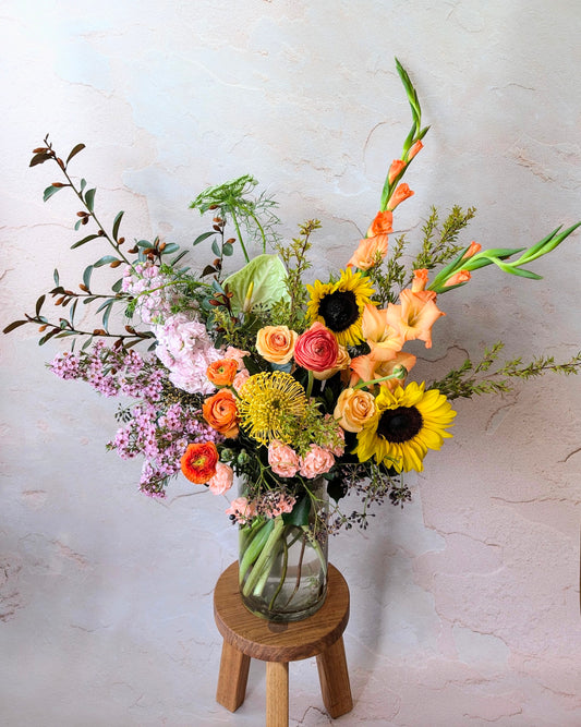 Vibrant floral arrangement with sunflowers, gladiolus, roses, and ranunculus in a clear vase on a wooden stool, handcrafted by Umasari Florist. Perfect for brightening home decor, events, and gifting. Available for delivery in Melbourne’s northern suburbs.