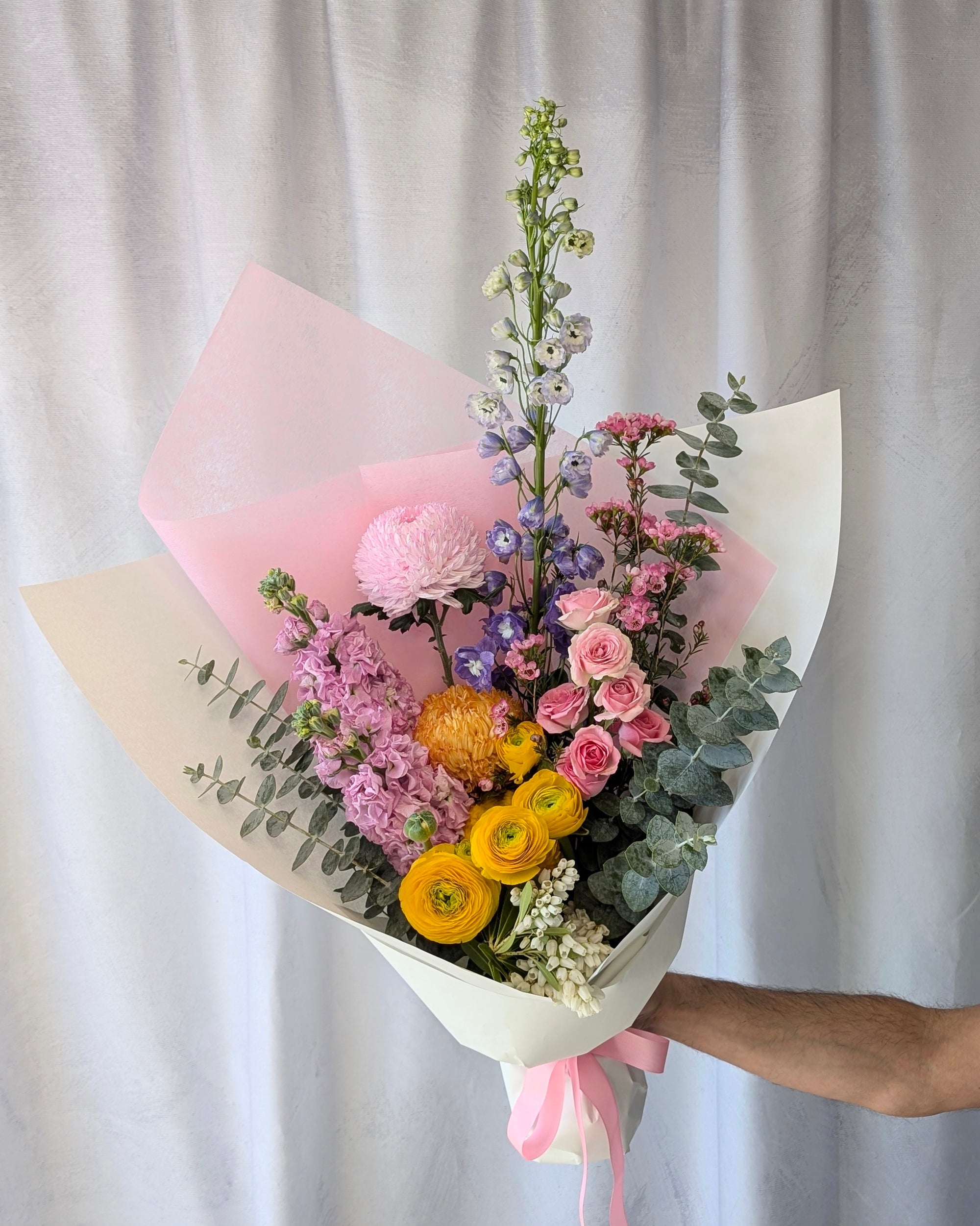 Bouquet of pink, yellow, purple and orange flowers wrapped in pink and white paper against a plain background. Available for flower delivery in Melbourne