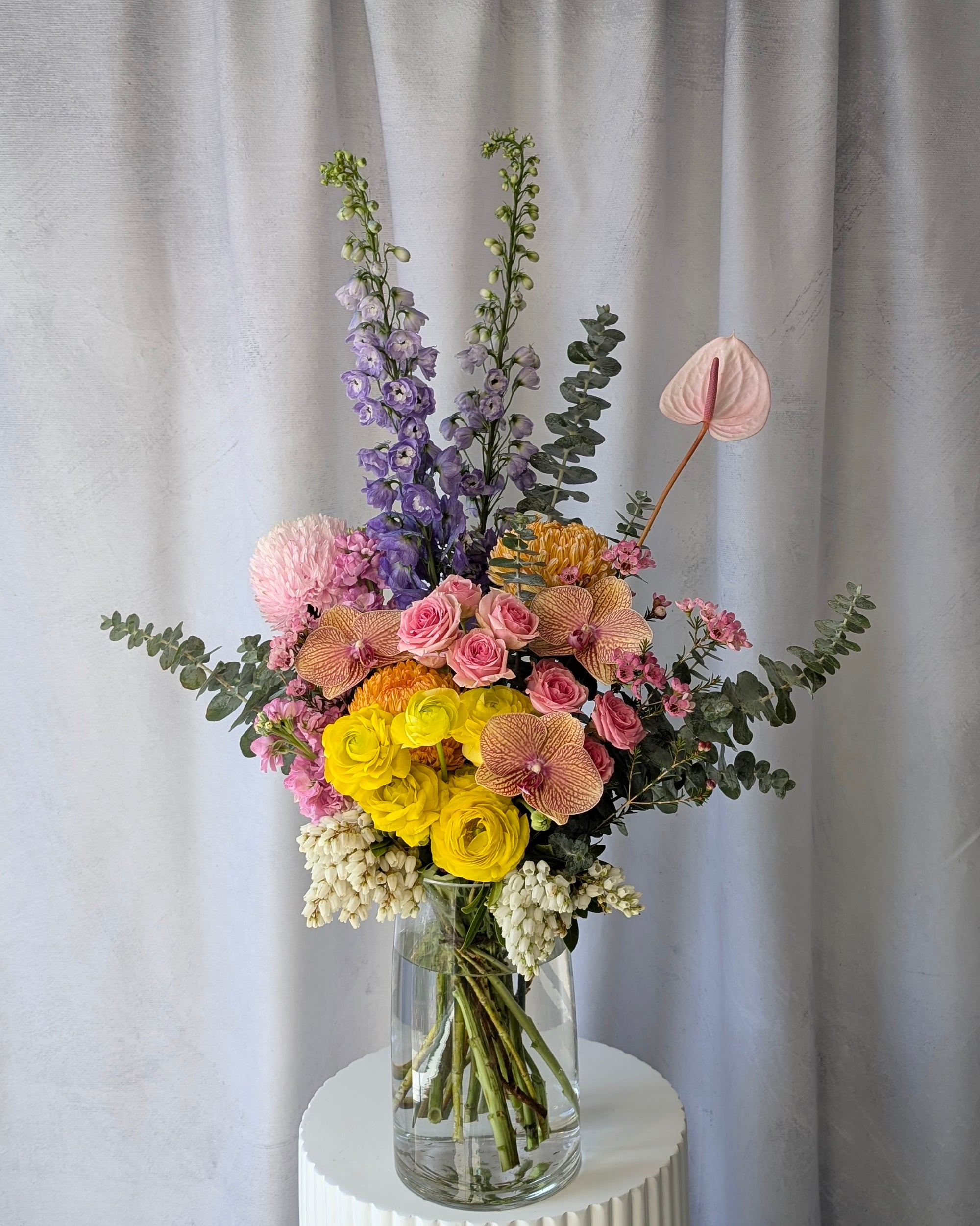 Colourful flower arrangement in a clear vase against a plain background. Flowers include delphiniums, anthuriums, ranunculus, roses and gum leaves. For delivery in Melbourne