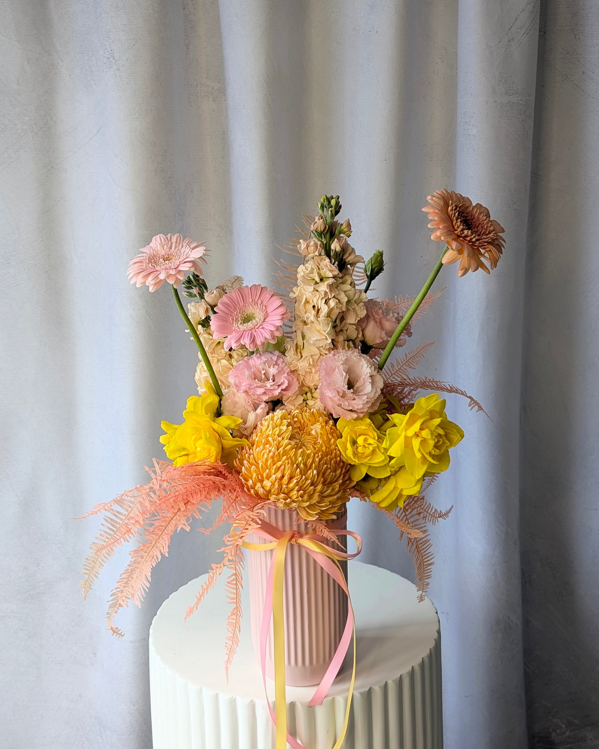 Colourful flower arrangement in a pink vase on a white surface with a light grey curtain background for delivery in Melbourne