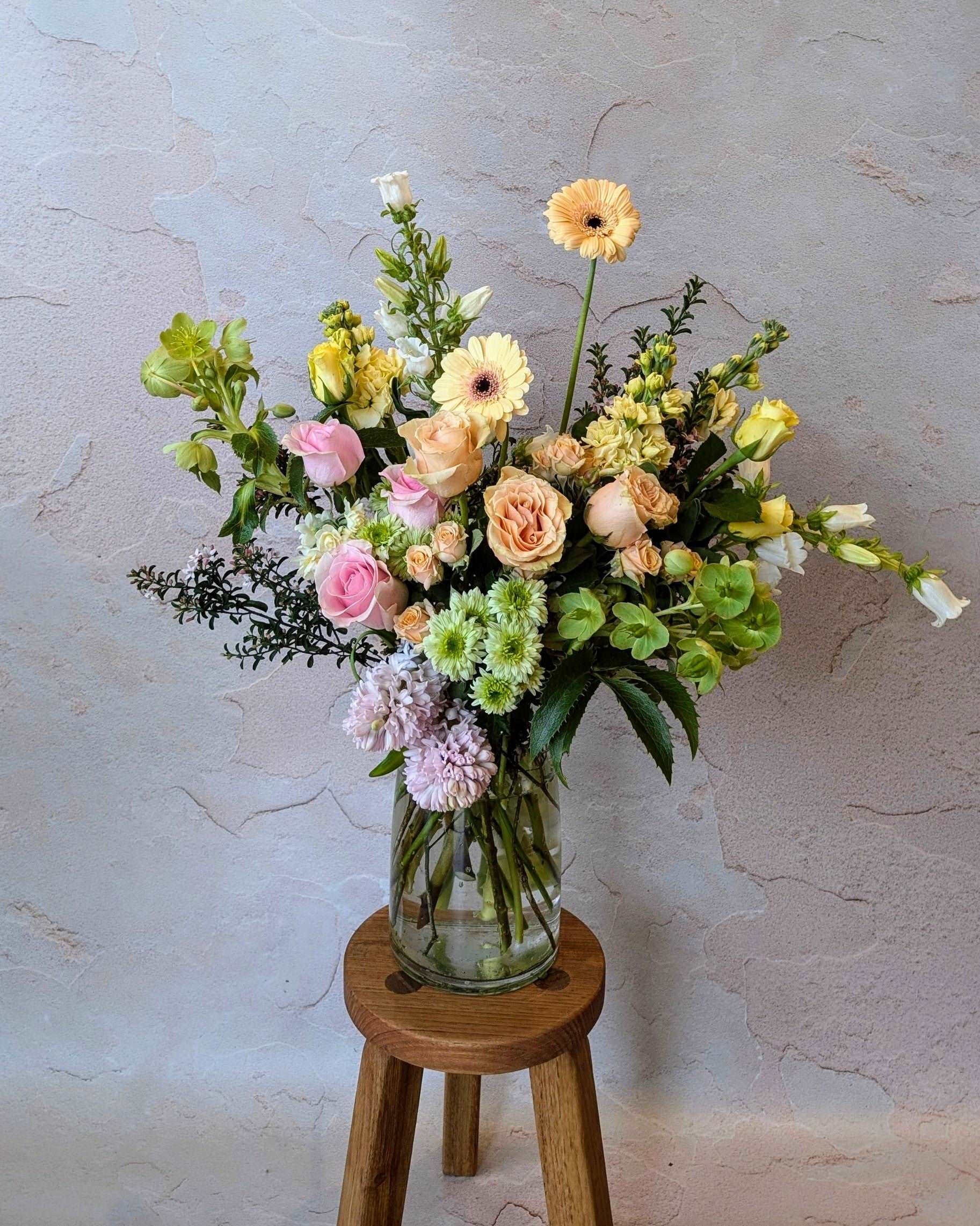 Elegant floral arrangement with soft pink roses, peach gerberas, chrysanthemums, and mixed greenery in a clear vase on a wooden stool. Perfect for weddings, home decor, or gift bouquets. The pastel shades and lush greenery create a fresh, romantic aesthetic