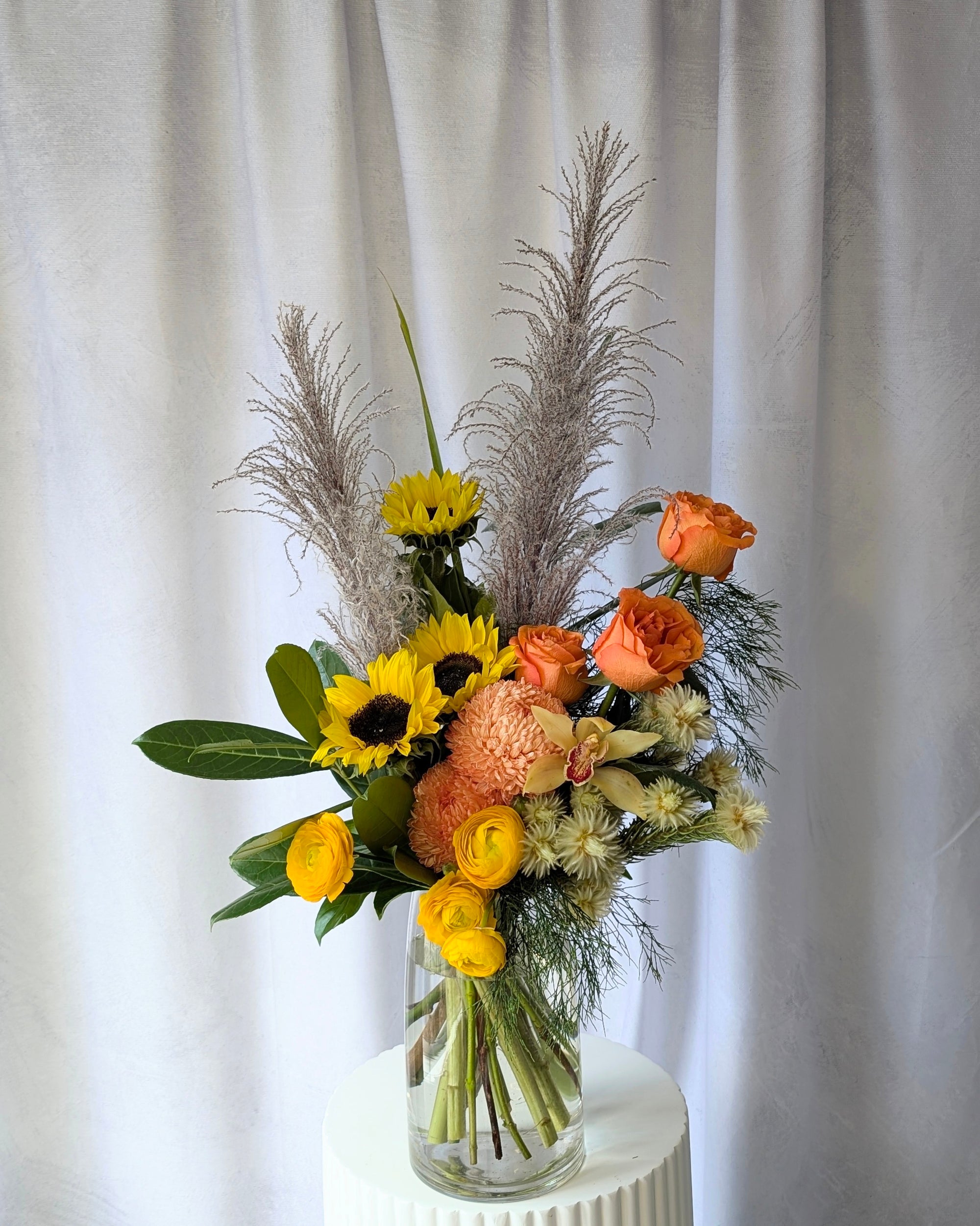 Bouquet of flowers with sunflowers, roses, and pampas grass in a clear vase against a white curtain background for delivery in Melbourne